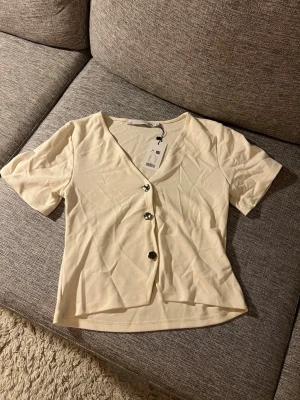 Vit v-ringad topp med knappar - Snygg vit topp med v-ringning och tre dekorativa knappar framtill. Kortärmad modell i mjukt material, perfekt till jeans eller kjol. Enkel och stilren design som passar till många olika looks.