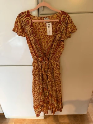 Blommig kortklänning från Spell - Supersöt lång klänning från Stajl med blommigt mönster i beige, rött och orange. Klänningen har v-ringning, volangdetaljer, knyt i midjan och korta ärmar. Perfekt för varma dagar och ger en bohemisk vibe.