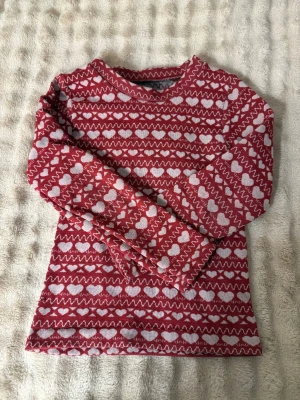 Röd stickad tröja med hjärtmönster - Mysig röd stickad tröja med vita hjärtan och zigzag-mönster. Långärmad och rund halsringning. Perfekt för kalla dagar och har en klassisk vinterkänsla. Materialet känns mjukt och värmande, troligen ullblandning. 