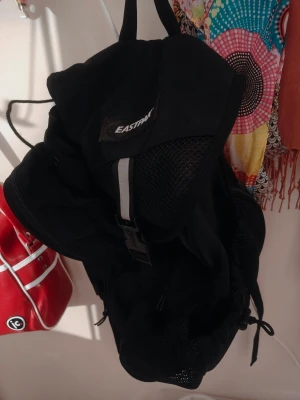 Svart ryggsäck från Eastpak Made In USA ( Nyskick) - Snygg svart ryggsäck från Eastpak med mesh-detaljer och flera fack. Väskan har justerbara axelband, dragkedjor i metall och spänne framtill. Perfekt för skolan eller utflykter, rymmer mycket och är enkel att bära.