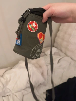 Grön Kipling handväska med patchar - Liten grön handväska från Kipling med flera färgglada patchar, bland annat ett hjärta och en Woodstock-lapp. Väskan har justerbar axelrem och dragskostängning upptill. Perfekt för dig som gillar en unik och personlig stil.