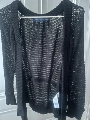 Svart stickad kofta French Connection - Svart, grovstickad kofta ( 72% bomull och 28 % ull) från French Connection med öppen front och ribbade muddar. Koftan har ett bälte i midjan för att knyta och är tillverkad i ett luftigt material som ger en avslappnad look. Perfekt för lager på lager och enkel att styla.