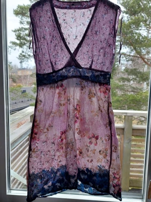 Blommigt transparent sommarklänning - Blommigt mönster i rosa, lila och blå toner. Djup V-ringning, dragkedja i sidan. Lätt och luftigt tyg, perfekt för varma dagar. .