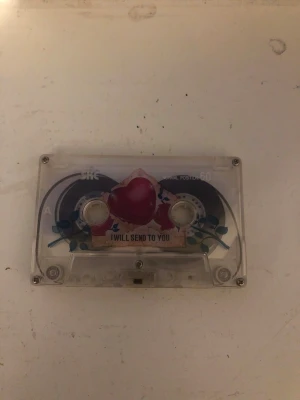 Genomskinligt kassettband SKC 60 - Säljer ett genomskinligt kassettband från SKC med kapacitet för 60 minuter. Kassetten har ett färgglatt klistermärke med ett rött hjärta och texten 'I WILL SEND TO YOU' på framsidan. Perfekt för dig som gillar retroprylar och vill spela eller samla på kassettband.