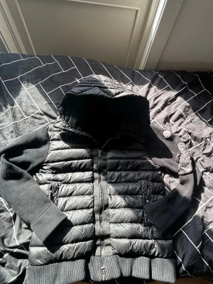 Svart cardiganjacka från Monclear Använd några gånger  - Snygg svart cardiganjacka från Monclear med quiltad kropp och stickade ärmar samt huva. Jackan har dragkedja framtill, ribbade muddar och två fickor med dragkedja. Perfekt för lager på lager och streetwear-stil. 