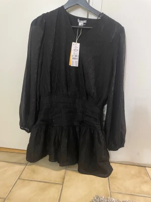 Svart omlottklänning med glitter - Snygg svart kortklänning med omlottdesign och subtilt glittrigt tyg. Klänningen har långärmade ärmar, markerad midja och lätt volang nedtill. Perfekt för dig som vill ha en trendig look med lite extra shine.