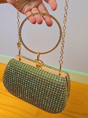 Glittrig kuvertväska med guldkedja - Säljer en superglittrig kuvertväska täckt av silvriga strass och med guldfärgade detaljer. Väskan har en rund guldfärgad handrem och en tunn guldkedja som axelband. Perfekt för dig som vill sticka ut och älskar bling! Materialet är hård metall och strass.