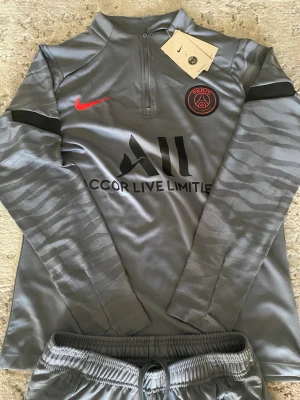 PSG träningsset i grått från Nike - Snyggt grått träningsset från Nike och Paris Saint-Germain med långärmad tröja och matchande byxor. Setet har svarta och röda detaljer, PSG-logga och Nike Swoosh. Materialet är glansigt och snabbtorkande, perfekt för fotbollsträning eller gym.