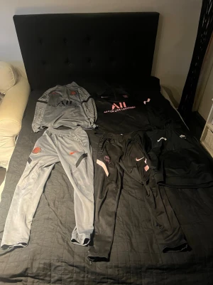 Nike PSG träningsset svart och grå - Säljer ett träningsset från Nike x PSG med två tröjor och två byxor. Ena setet är grått med orange PSG-logga och Nike swoosh, det andra är svart med rosa detaljer och PSG-märke. Dresserna är i storlek S medans Nike air tröjan är i storlek L i barn, 147-158cm.Materialet är funktionspolyester, perfekt för träning eller chill. Snygg design med dragkedja på tröjan.