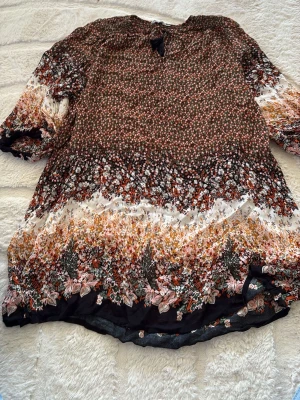 Blommig blus från Indiska - Mönstrad blus från Indiska i storlek XL med småblommigt tryck i grönt, orange, vitt och svart. Blusen har trekvartsärm och rund hals med knytband. Tillverkad i ett lätt och luftigt tyg som ger en bohemisk känsla.