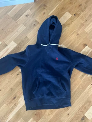Mörkblå hoodie från Polo Ralph Lauren - Mörkblå hoodie från Polo Ralph Lauren med klassisk huva och vita dragsnören. Har en känguruficka framtill och den ikoniska röda logotypen broderad på bröstet. Perfekt för en avslappnad och stilren look. Tillverkad i mjukt bomullsmaterial. Nyskick inte använt alls mycket