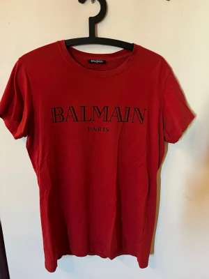 Röd Balmain Paris t-shirt - Snygg röd t-shirt från Balmain Paris med klassisk logga tryckt framtill. T-shirten har rund halsringning och korta ärmar. Tillverkad i mjuk bomull som känns skön mot huden. Perfekt för dig som vill ha en stilren och trendig look.