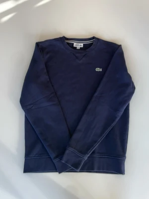 Mörkblå sweatshirt från Lacoste Sport - Mörkblå sweatshirt från Lacoste Sport med klassisk rund halsringning och det ikoniska krokodilmärket broderat på bröstet. Tröjan har långa ärmar och ribbade muddar vid ärmslut och nederkant. Perfekt för en avslappnad och sportig stil.