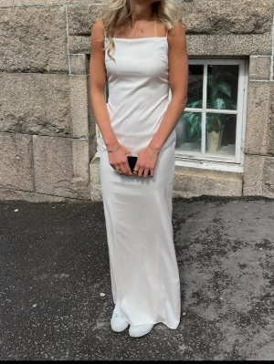 Ljusrosa balklänning - Elegant vit långklänning med smala axelband och rak halsringning. Klänningen har en slät, minimalistisk design och är tillverkad i ett glansigt material som ger en lyxig känsla. Perfekt för student eller sommarens speciella tillfällen. Balklänning