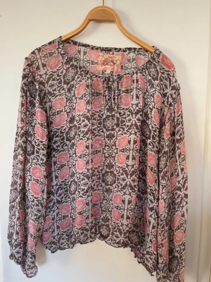 Mönstrad blus med rosa detaljer - Säljer en luftig blus med lång ärm och rund halsringning. Blusen har ett bohemiskt mönster i svart och rosa på transparent tyg. Perfekt för dig som gillar en avslappnad och trendig stil.