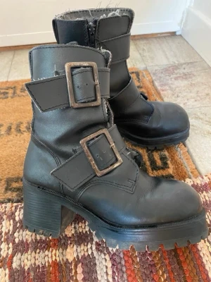 Svarta chunky stövletter med spännen - Tuffa svarta stövletter i skinn med grov sula och klack. Två stora metallspännen ger en edgy look och dragkedja på insidan gör dem enkla att ta på. Perfekta för dig som gillar en rockig stil.