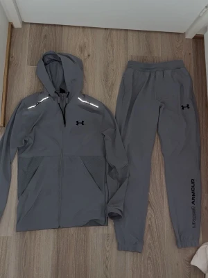 Grå träningsset från Under Armour - Snyggt grått träningsset från Under Armour med huva och dragkedja på jackan samt matchande byxor. Setet har reflekterande detaljer på axlarna och logga på både jacka och byxa. Perfekt för löpning eller gym. Tillverkat i lätt och funktionellt material.
