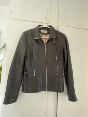 Brun skinnjacka från Biba - Snygg mörkbrun skinnjacka från Biba med raka dragkedjor framtill och två fickor med dragkedja. Jackan har ståkrage med tryckknapp och coola sömdetaljer vid sidorna. Fodret är randigt i orange och beige toner för en extra touch.