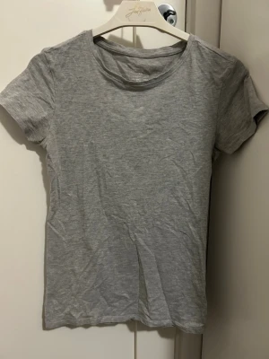 Grå basic t-shirt från Primark - En enkel och stilren grå t-shirt från Primark med rund halsringning och korta ärmar. Tillverkad i mjuk bomull med stretch för extra komfort. Perfekt att matcha med jeans eller shorts för en avslappnad look. Tröjan är i storlek XS.