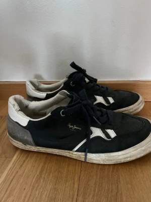 Svarta låga sneakers från Pepe Jeans - Säljer ett par svarta sneakers från Pepe Jeans med vita detaljer och snörning. Skorna har en klassisk låg siluett och är tillverkade i mocka och textil. Pepe Jeans-loggan syns på sidan och sulan är vit med en något sliten look.
