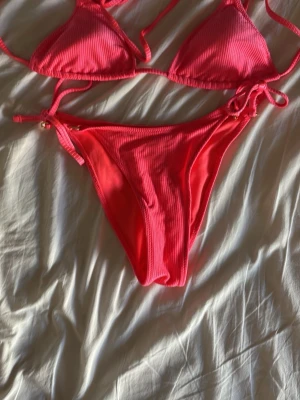 Rosa ribbad trekantsbikini - Säljer en snygg rosa bikini med ribbad struktur. Överdelen är trekantsmodell med knyt i nacken och ryggen, och underdelen har knyt i sidorna. 