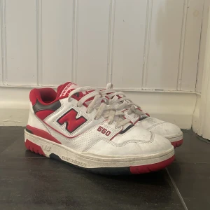 New Balance 550 sneakers i vitt och rött - Snygga New Balance 550 sneakers i vitt med röda och svarta detaljer. Skorna har snörning och en klassisk design med märkets logga på sidan och hälen. Perfekta för en sportig look.