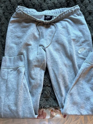 Gråa mjukisbyxor från Nike - Snygga ljusgrå mjukisbyxor från Nike med dragsko i midjan och resår vid bensluten. Byxorna har en broderad Nike-logga på vänster ben och är gjorda i mjukt bomullsmaterial. Perfekta för chill dagar eller träning.