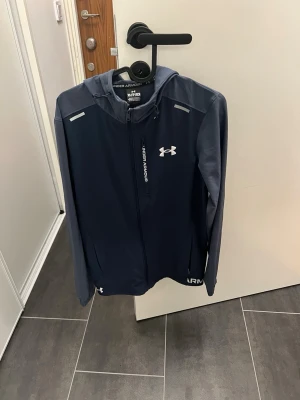 Blå träningsjacka från Under Armour - Snygg marinblå träningsjacka från Under Armour med huva och hel dragkedja. Jackan har vita logotyper på bröstet och dragkedjan, samt reflekterande detaljer på axlarna. Tillverkad i lätt och tekniskt material som passar perfekt för löpning eller gym. Använd 1 gång bara köpt för 2700
