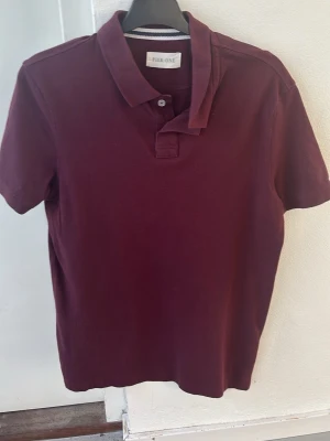 Vinröd pikétröja från Pier One - Snygg vinröd pikétröja från Pier One med klassisk krage och knappslå. Tröjan har korta ärmar och är tillverkad i mjukt bomullsmaterial. Perfekt för en avslappnad men stilren look. Storlek S men passar även M