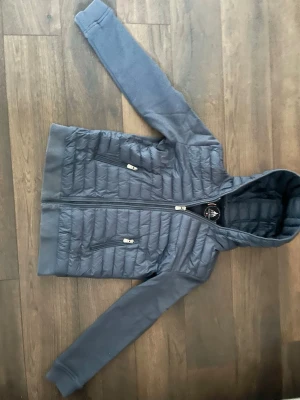 Blå pufferjacka med huva från JOTT - Snygg blå pufferjacka från JOTT med huva och dragkedja framtill. Jackan har quiltad kropp, mjuka ärmar och två dragkedjeförsedda fickor. Perfekt för kalla vinterdagar och har en modern, sportig look.