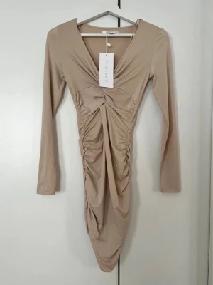 Beige omlottklänning från Amisu - Trendig beige klänning från Amisu med omlottdesign och rynkade detaljer framtill. Klänningen har lång ärm, v-ringning och är tillverkad i ett mjukt, stretchigt material som ger en smickrande passform.