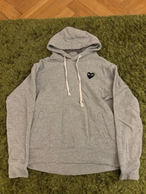 Grå hoodie Comme des Garçons Play - Säljer en grå hoodie från Comme des Garçons Play med det ikoniska svarta hjärtat med ögon broderat på bröstet. Hoodien har inga defekter eller sådant och är i storlek S. Hör av vid funderingar