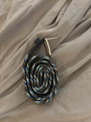 Randigt flätat bälte i blå toner - Snyggt flätat bälte med randigt mönster i blått, svart, vitt och brunt. Bältet har ett silvrigt spänne och detaljer i svart skinn. Perfekt accessoar för att lyfta din outfit.