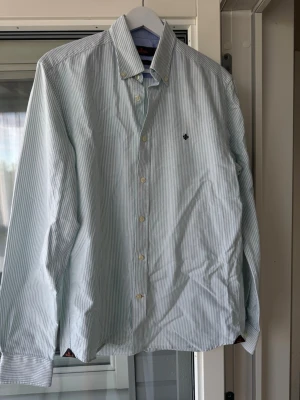 Randig skjorta från Morris - Snygg ljusblå och vit randig skjorta från Morris med klassisk button-down krage och broderad logga på bröstet. Skjortan har långa ärmar och knäppning framtill. Perfekt för en stilren look.