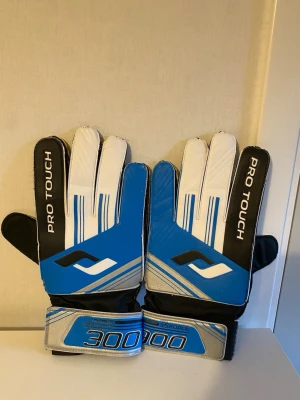 Blåa målvaktshandskar Pro Touch - Snygga målvaktshandskar från Pro Touch i blått, vitt och svart med silverdetaljer och justerbar kardborre vid handleden. Perfekta för fotbollsträning eller match. Modell 3000 med greppvänlig insida och sportig design.