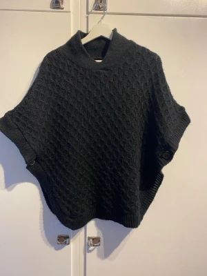 Svart stickad poncho med polokrage - Snygg svart stickad poncho med polokrage och korta ärmar. Ponchon har ett diskret rutmönster och ribbstickade kanter. Har ingen storlek, men sitter bra på mig med s-m