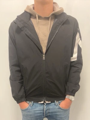Svart vindjacka från Moncler - Svart vindjacka från Moncler med vit M-detalj på ärmen och dragkedja framtill. Jackan har huva, elastiska muddar och två sidofickor. Perfekt för en sportig och stilren look.