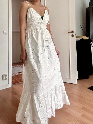 Vit långklänning med broderade detaljer - Säljer en vit långklänning med tunna axelband och v-ringning. Klänningen har broderade blommönster, smock i midjan och knyt framtill. Perfekt för sommarens varma dagar och har en luftig, romantisk känsla.