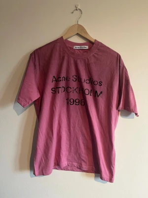 Rosa Acne Studios t-shirt Stockholm - Säljer en rosa t-shirt från Acne Studios med svart tryck framtill där det står 'Acne Studios STOCKHOLM 1996'. Klassisk rund hals och korta ärmar. Tillverkad i mjuk bomull för en skön och avslappnad passform. 