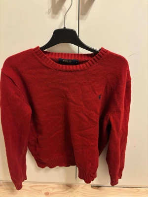 Röd stickad tröja Polo Ralph Lauren - Röd stickad tröja från Polo Ralph Lauren med rund halsringning och ribbade muddar vid ärmslut och nederkant. Klassisk liten blå logga broderad på bröstet. Perfekt för dig som gillar stilrena och tidlösa plagg.