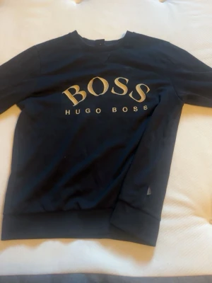 Svart sweatshirt från Hugo Boss - Snygg svart sweatshirt från Hugo Boss med stort BOSS-tryck i beige på bröstet. Klassisk rund halsringning och ribbade muddar vid ärmslut och nederkant. Perfekt för en stilren och avslappnad look.