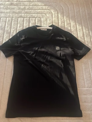 Svart Stone Island t-shirt - Snygg svart t-shirt från Stone Island med klassisk logopatch på bröstet. T-shirten har rund hals och korta ärmar, tillverkad i mjuk bomull för en skön känsla. Perfekt för dig som gillar stilrena och ikoniska plagg.