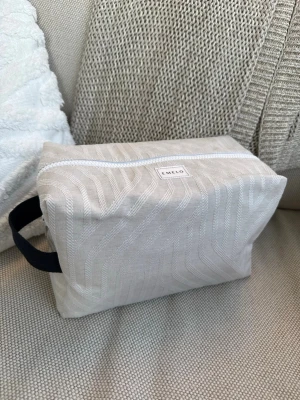 Ljusbeige necessär från Emelo - Snygg ljusbeige necessär från Emelo med subtilt mönster i tyget. Rektangulär form med vit dragkedja och svart handtag på sidan. Perfekt för att organisera smink eller småprylar. Tillverkad i textilmaterial.