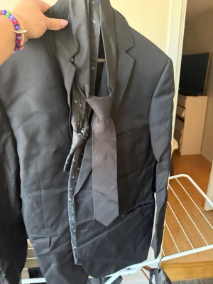 Svart kostymkavaj med slips - Svart kostymkavaj med klassisk passform och matchande svart slips. Kavajen har enkla slag och två fickor framtill. Perfekt för dig som vill ha en stilren och enkel look. Materialet ser ut att vara polyester eller liknande.