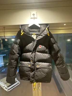 Svart jacka från Fjällräven - Säljer en svart dunjacka från Fjällräven. Jackan har en stor huva, två stora fickor framtill och klassisk Fjällräven-logga på bröstet. Perfekt för kalla vinterdagar och riktigt varm tack vare dunfyllningen. Använd ett fåtal gånger                                             Kan gå ner i pris vid snabb affär. Köpt för: 8300
