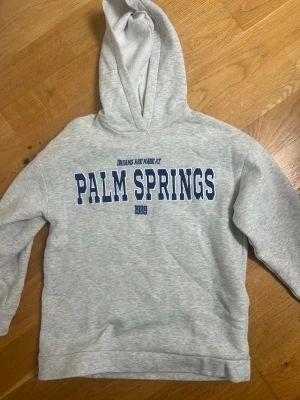Grå hoodie Palm Springs Gina Tricot - Snygg ljusgrå hoodie från Gina Tricot med blå text 'PALM SPRINGS' och 'Dreams are made at' framtill. Mjuk insida och klassisk huva. Perfekt för chill dagar och streetstyle. Tillverkad i bomullsblandning för extra komfort.