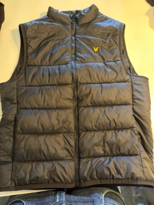 Svart dunväst från Lyle & Scott - Snygg svart dunväst från Lyle & Scott med gul broderad logga på bröstet. Västen har hög krage, dragkedja framtill och quiltad design med breda paneler. Perfekt att slänga över en hoodie eller tröja för extra värme.