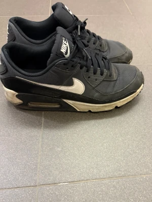 Svarta Nike Air Max sneakers - Snygga svarta Nike Air Max sneakers med vit sula och klassisk vit swoosh på sidan. Skorna har en sportig design med mesh och syntetmaterial, samt synlig Air-dämpning i sulan för extra komfort. Perfekta för dig som gillar streetwear och vill ha en ikonisk look.