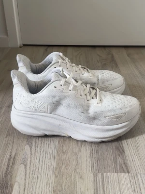 Vita Hoka sneakers med tjock sula - Säljer ett par helvita Hoka sneakers med riktigt chunky och dämpad sula. Ovandelen är i meshmaterial som andas och har snygga detaljer samt logga på sidan. Skorna har klassisk snörning och en sportig, modern look.