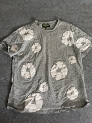 Grå t-shirt med vita blommor - Grå t-shirt från Green House med vita blommönster utspridda över hela tröjan. Klassisk rund halsringning och korta ärmar. Perfekt för en avslappnad och trendig stil.